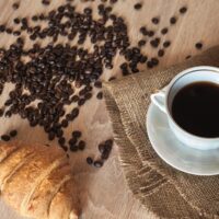 French Roast Coffee. A cup of fragrant black coffee on a saucer, a croissant and coffee beans on a wooden brown textured table covered with linen material.