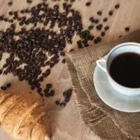 French Roast Coffee. A cup of fragrant black coffee on a saucer, a croissant and coffee beans on a wooden brown textured table covered with linen material.